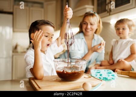La giovane madre e i suoi figli gustano la pasta fresca con cioccolato fuso. Donna e bambini cucinano in cucina, preparazione di torte. Buona famiglia Foto Stock