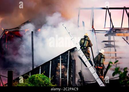i vigili del fuoco combattono contro un fuoco selvatico sullo sfondo di un fumo bianco spesso. I vigili del fuoco sulle scale spengono il tetto di una casa privata o fienile. Foto Stock