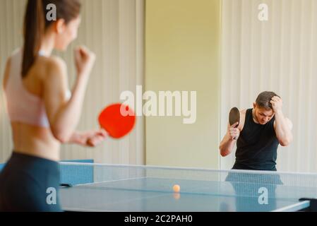 Uomo e donna che giocano a ping pong al chiuso, ragazza vincitore. Coppia in abbigliamento sportivo tiene racchette e gioca a ping pong in palestra Foto Stock