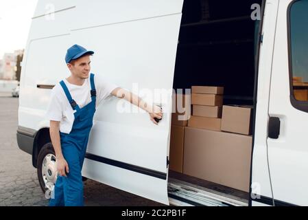 Consegna in porta auto a chiusura uniforme, auto con pacchi e scatole di cartone, servizio di consegna. Uomo in piedi a confezioni di cartone in veicolo, maschio del Foto Stock