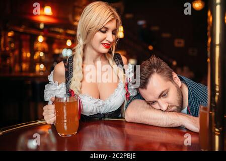 Bella cameriera con boccale di birra si affaccia su uomo ubriaco al bancone in pub, oktoberfest vacanze. Persona di sesso maschile di bere in bar, barista in tradizionale retr Foto Stock