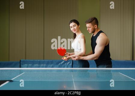 L'uomo insegna a una donna a giocare a ping pong, allenarsi in interni. Coppia in abbigliamento sportivo tiene racchette e gioca a ping pong in palestra Foto Stock