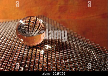 Griglia metallica riflessa in lensball trasparente. Sfera di vetro che giace sulla griglia contro sfondo arancione. Foto Stock
