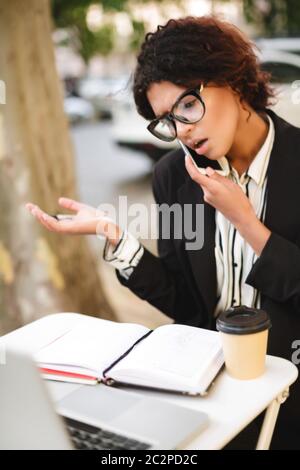Ritratto di afroamericana ragazza in occhiali seduti al tavolo e emotivamente parlando sul suo cellulare. Donna con capelli ricci scuri e dal aspetto sorprendente Foto Stock