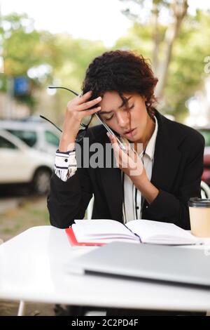 Ritratto di afroamericana seduta al tavolo con gli occhiali in mano e chiudendo gli occhi stancamente mentre parla sul cellulare. Donna con cu scuro Foto Stock