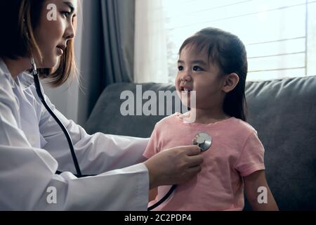 La bambina asiatica sorride e si siede sul divano a casa durante un esame medico mentre la donna professionista sta usando uno stetoscopio per liste Foto Stock