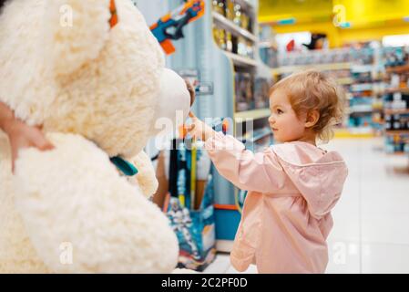 Bambina che sceglie l'orso grande del teddy nel deposito dei capretti, vista laterale. Figlia in cerca di giocattoli in supermercato, shopping di famiglia, giovane cliente Foto Stock