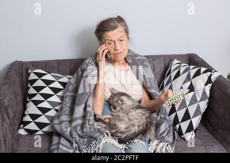 Donna anziana malata che prende la medicina a casa. Donna anziana che parla sul telefono cellulare mentre guarda la medicina in camera da letto. Pensionato Foto Stock