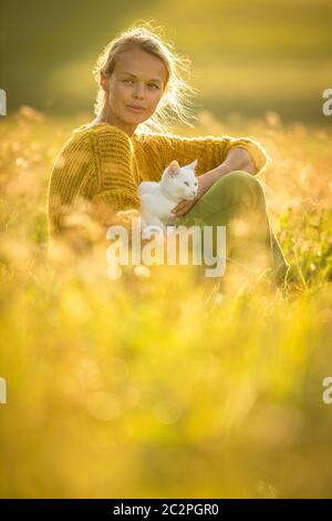 Piuttosto, giovane donna con il suo gatto Pet sitting in erba sul bellissimo prato illuminato dalla calda luce della sera (SHALLOW DOF; dai toni di colore immagine) Foto Stock