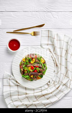 Dieta di manzo al keto con broccoli, fagioli verdi, peperoni dolci gialli e rossi, prezzemolo, cipolla arrostita su un piatto bianco su fondo bianco di legno, Foto Stock