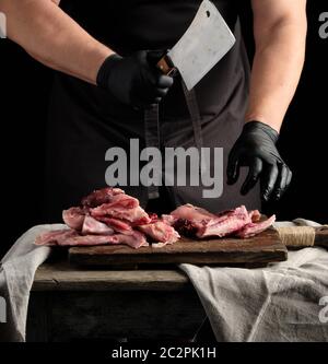 Chef in nero Guanti in lattice tiene un coltello di grosse dimensioni e tagli in pezzi grezzi di carne di coniglio su un marrone tagliere di legno, cottura su uno sfondo scuro Foto Stock