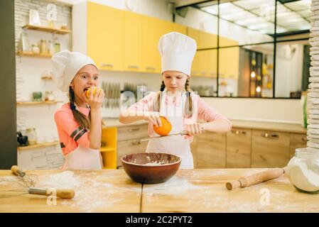 Due bambine cuoce in caps strofina l'arancia alla ciotola, preparazione dei biscotti sulla cucina. Pasticceria per bambini, chef per bambini che preparano la torta Foto Stock