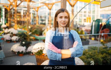 Donna giardiniere pone in negozio per giardinaggio, venditore. Donna vende piante in negozio fiorista, venditore in grembiule e guanti Foto Stock