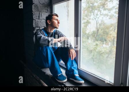 Uomo depresso seduto sul davanzale della finestra in camera oscura, psycho paziente. Concetto di persone mentalmente malate, umano sottolineato, depressione Foto Stock