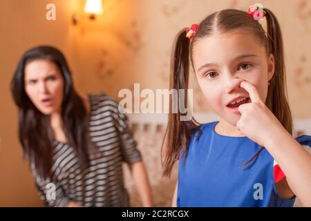 Ragazza curiosa che raccoglie un naso contro la madre sorpresa sullo sfondo. Foto Stock