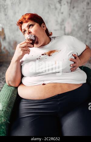 Il sovrappeso donna si siede su una sedia e mangia la torta dolce, la pigrizia e l'obesità, bulimic. Cibo malsano mangiare, femmina grassi Foto Stock