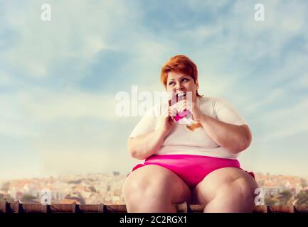 Il sovrappeso donna si siede sul tetto e mangia la torta dolce, la pigrizia e l'obesità, bulimic. Cibo malsano mangiare, femmina grassi Foto Stock