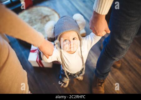 Divertente bambino ragazzo piccolo 1 anno di apprendimento camminare a casa in inverno in una casa di Capodanno decorato. Giovane famiglia papà e mamma tenere da Th Foto Stock