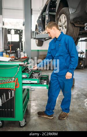 Repairman raggiungendo per la chiave nella cassetta degli attrezzi, auto sul ponte sullo sfondo. Automobile equipaggiamenti di servizio e di manutenzione del veicolo Foto Stock