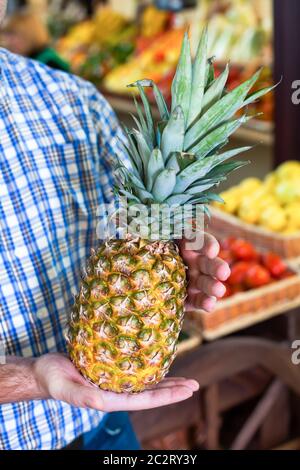 Ritratto di uomo sorridente in camicia tiene l'ananas maturo in mani. Negozio di alimentari sullo sfondo. Foto Stock