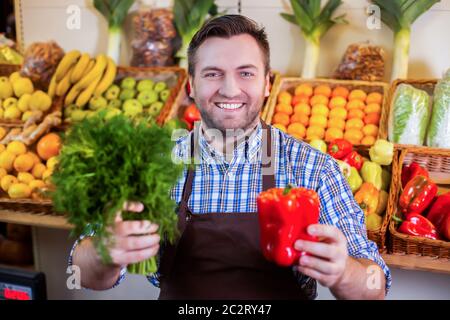 Venditore sorridente in grembiule che mostra verdure fresche al negozio di grossery. Scatole con frutta e verdura sullo sfondo. Foto Stock