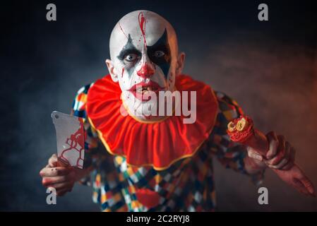 Mad sanguinosa clown con carne cleaver e mano umana. L'uomo con il trucco in costume di halloween, crazy maniac, horror Foto Stock