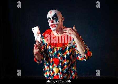 Mad sanguinosa clown con carne cleaver, circus orrore. L'uomo con il trucco in costume di carnevale, crazy maniac Foto Stock