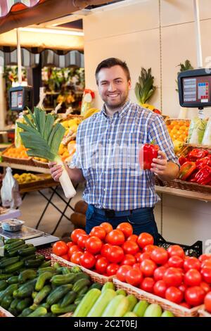 Il venditore sorridente mostra le verdure in negozio. Scatole con frutta e verdura sullo sfondo. Foto Stock