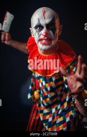 Mad sanguinosa clown con carne cleaver e la mazza da baseball, circus orrore. L'uomo con il trucco in costume di carnevale Foto Stock