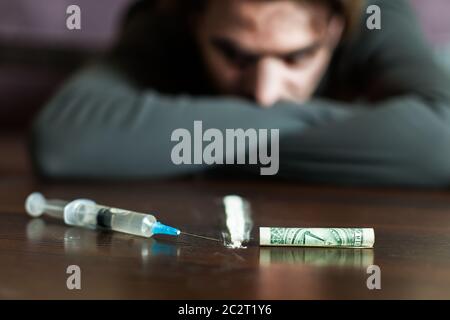 L'uomo di dipendenza guarda il tavolo con siringa, dollari e traccia di cocaina. L'abuso di stupefacenti porta ad una depressione. Foto Stock