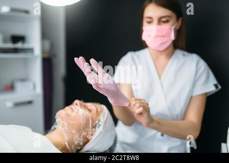 Estetista indossa guanti contro il paziente con la maschera per il viso, sbarazzarsi di rughe, cosmetologia clinica. Cura della pelle del viso, procedura di ringiovanimento nella spa sa Foto Stock