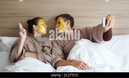 la giovane famiglia con accappatoi e maschere dorate fa selfie con smartphone sdraiato in un letto morbido in una comoda camera d'hotel Foto Stock