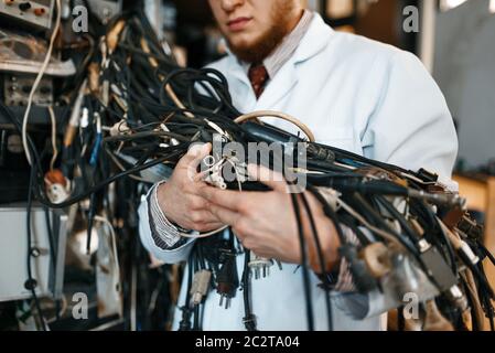 Un ingegnere strano può tenere in laboratorio un gruppo di fili con diversi connettori. Strumenti di prova elettrica in background. Attrezzature da laboratorio, officina di ingegneria Foto Stock