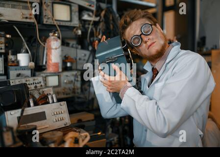 Crazy scienziato in occhiali contiene dispositivi elettrici in laboratorio. Strumenti di prova elettrica in background. Attrezzature da laboratorio, officina di ingegneria Foto Stock