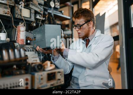 Crazy scienziato in occhiali contiene dispositivi elettrici in laboratorio. Strumenti di prova elettrica in background. Apparecchiature da laboratorio Foto Stock