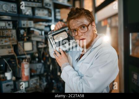 Crazy scienziato in occhiali contiene dispositivi elettrici in laboratorio. Strumenti di prova elettrica in background. Apparecchiature da laboratorio Foto Stock