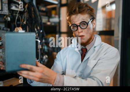 Crazy scienziato in occhiali contiene dispositivi elettrici in laboratorio. Strumenti di prova elettrica in background. Attrezzature da laboratorio, officina di ingegneria Foto Stock