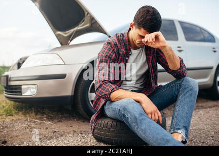 Stanco uomo seduto sul pneumatico, Broken auto con cappa aperta sullo sfondo Foto Stock