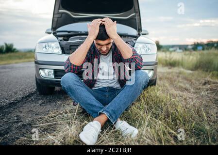 Stanco uomo seduto sul pneumatico, Broken auto con cappa aperta sullo sfondo Foto Stock