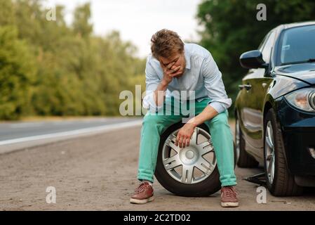 Guasto dell'auto, uomo stanco seduto sulla ruota di scorta. Auto rotta o problema con il veicolo, problema con pneumatico forato auto su strada Foto Stock