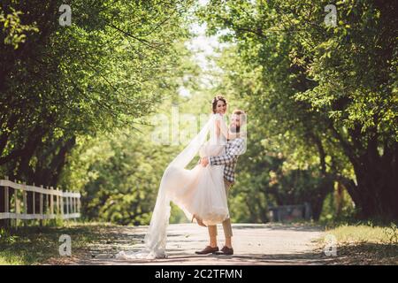 Felice sposo alza sposa nel parco estivo. Uomo alzato e abbraccio donna alle braccia. Coppia appena sposata nel parco. Solo matrimonio Foto Stock