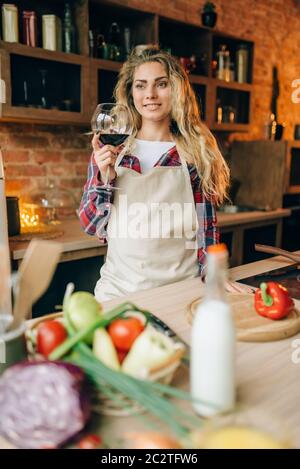 Felice casalinga in un grembiule tenendo un bicchiere di vino rosso nelle mani, cucina interno su sfondo. Cuoca prima di insalata di verdure preparazione Foto Stock