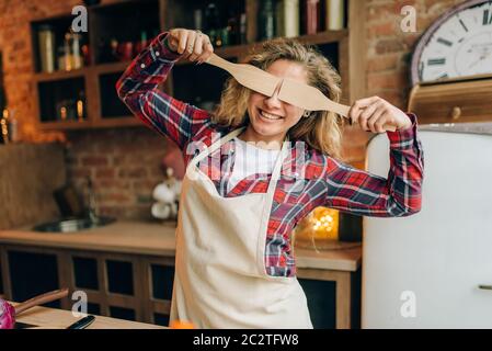 Giocoso casalinga in un grembiule copre gli occhi con le spatole in legno, cucina interno su sfondo. Felice cuoca facendo una sana alimentazione vegetariana, Foto Stock