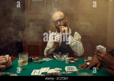 Giocatore di poker con bearded con sigaro, casinò. Giochi di dipendenza casuale. Uomo svaghi in casa gioco d'azzardo. Carte, patatine e whisky sul tavolo da gioco Foto Stock