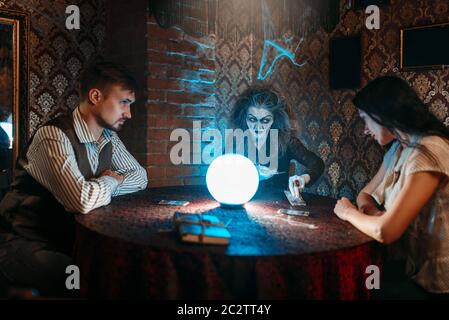 Strega legge una magia incantesimo su una sfera di cristallo, una giovane coppia in seance spirituale. Femmina chiamate foreteller gli spiriti Foto Stock