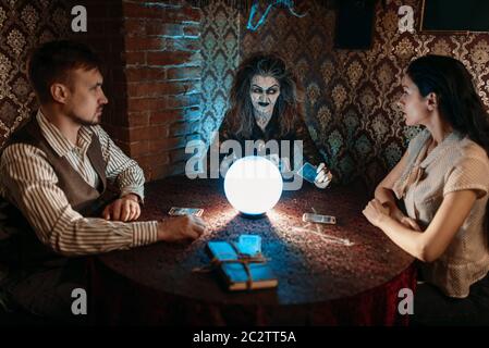 Scary witch legge una magia incantesimo su una sfera di cristallo, giovane uomo e donna su seance spirituale. Femmina chiamate foreteller gli spiriti Foto Stock