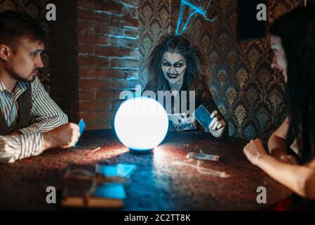 Strega legge una magia incantesimo su una sfera di cristallo, una giovane coppia in seance spirituale. Femmina chiamate foreteller gli spiriti Foto Stock