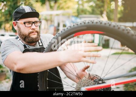 Noleggio repairman funziona con la ruota di bicicletta, ciclo workshop all'aperto. Barbuto meccanico nel grembiule Foto Stock