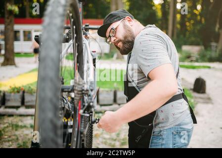 Repairman funziona con la ruota di bicicletta, ciclo workshop all'aperto. Bicicletta barbuto meccanico nel grembiule Foto Stock