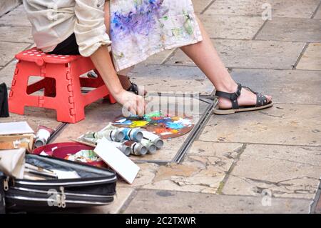 Donna artista pittura sulla strada, è seduta su una panca rossa, attrezzi dipingono accanto ai piedi. Foto Stock